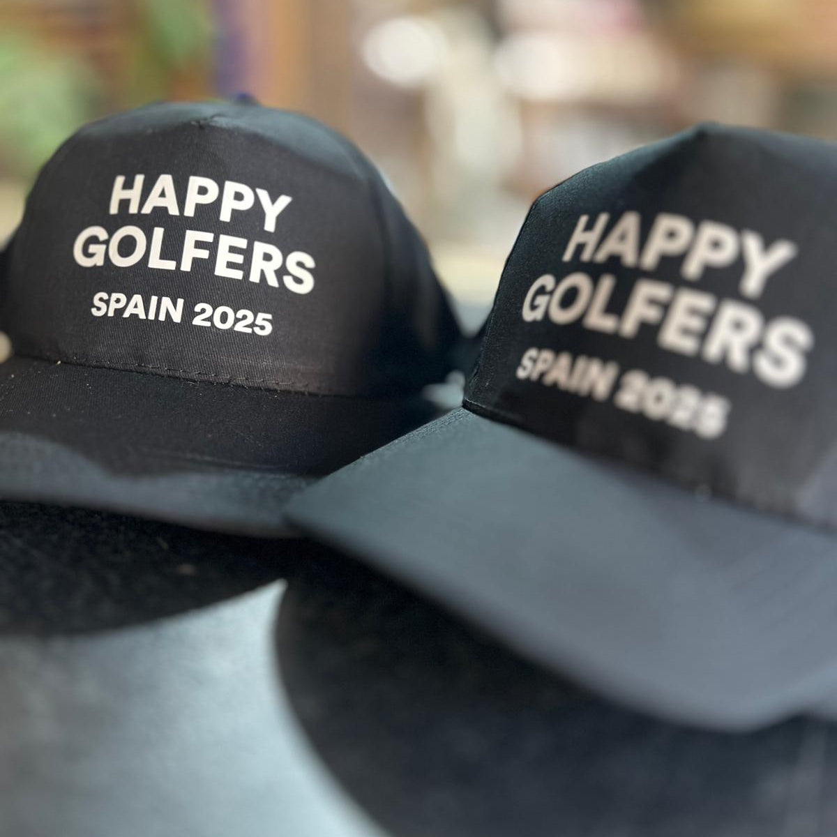Two black caps printed with 'Happy Golfers Spain 2025' text on a blurred background. Made by The Wrong End of Town in Dover, Kent.